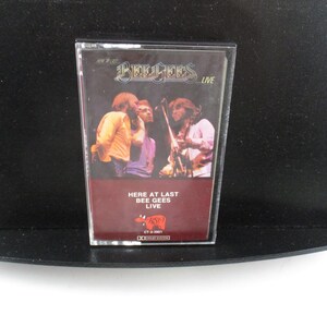 May include: A cassette tape case with a picture of the Bee Gees band members on the front. The case is red and white with the text "Here at Last Bee Gees Live" printed on the front.