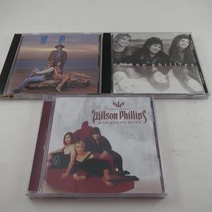 May include: Three Wilson Phillips music CDs. The top left CD features a man and two women on the cover. The top right CD has a black and white photo of three women. The bottom CD is labeled "Greatest Hits" and features four women.