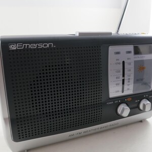 May include: Black and silver AM/FM weather band radio with the Emerson logo. The radio has a speaker grill, a tuning dial, and a digital clock display. The words "AM/FM WEATHER BAND RADIO" are printed on the bottom.