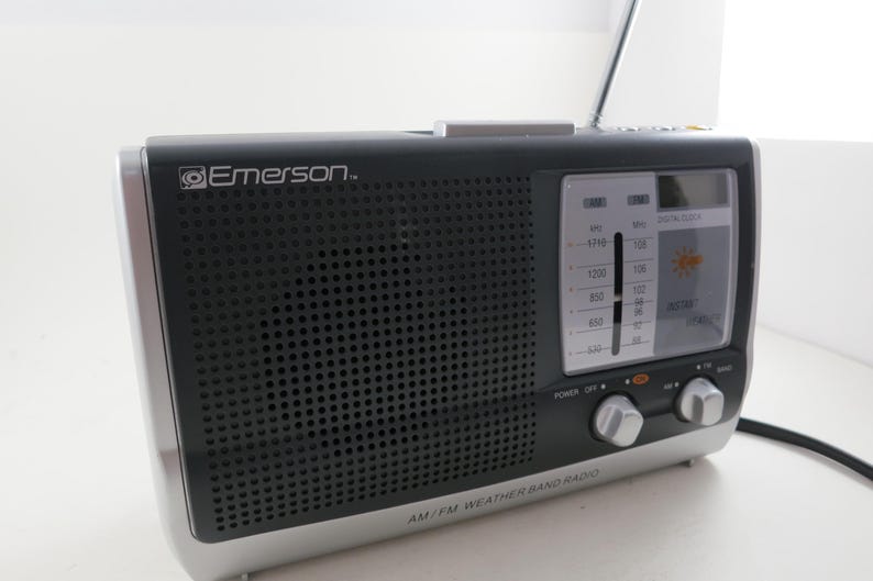 May include: Black and silver AM/FM weather band radio with the Emerson logo. The radio has a speaker grill, a tuning dial, and a digital clock display. The words "AM/FM WEATHER BAND RADIO" are printed on the bottom.