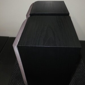 May include: Two black speakers with a wood grain finish. The speakers have a unique angled design with a silver trim. The speakers are likely for a home entertainment system.