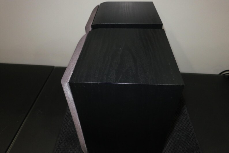 May include: Two black speakers with a wood grain finish. The speakers have a unique angled design with a silver trim. The speakers are likely for a home entertainment system.