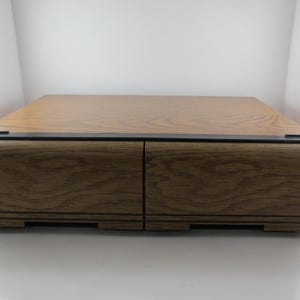 May include: A wooden storage box with two drawers. The box has a light brown top and a darker brown, wood-grain finish on the drawers and sides. The top has a black trim. The drawers are closed.