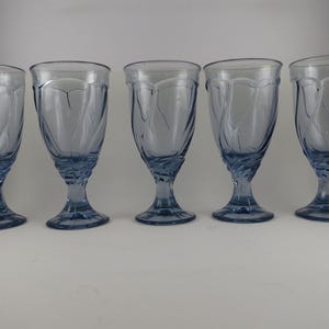 Pu&ograve; includere: Cinque calici d'acqua bluastri con un design decorativo a spirale sulla coppa e sullo stelo. I bicchieri sono di vetro e sono disposti in fila, mostrando la loro forma e il loro colore eleganti.