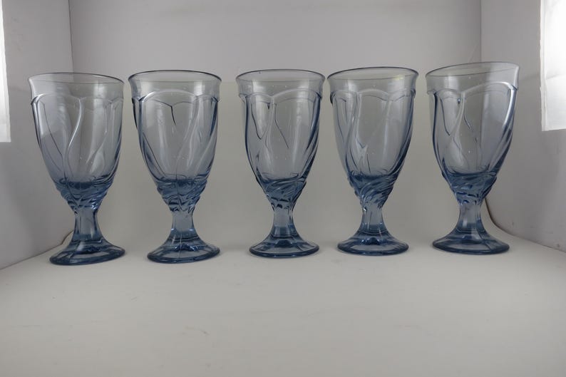 Pu&ograve; includere: Cinque calici d'acqua bluastri con un design decorativo a spirale sulla coppa e sullo stelo. I bicchieri sono di vetro e sono disposti in fila, mostrando la loro forma e il loro colore eleganti.