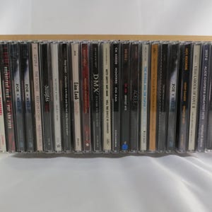 May include: A collection of twenty-six music CDs arranged side-by-side in a row. Each CD case features a thin spine with printed text displaying various artist names and album titles, including works by Adele, DMX, and Alice Cooper in a rack.