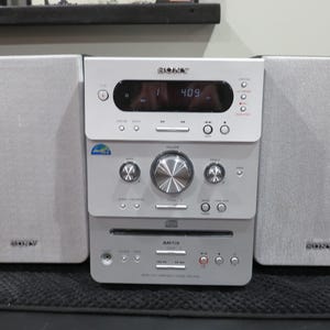 May include: A silver Sony stereo system with two speakers. The stereo has a CD player, radio, and display screen showing the time. The speakers and stereo are silver and have the Sony logo.