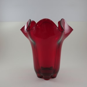May include: A vibrant red glass vase with a ruffled rim. The vase has a flared, tulip-like shape and a smooth, glossy finish. The base has a unique, three-footed design. A decorative piece for the home.