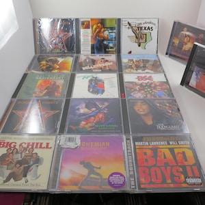 May include: A collection of 16 compact disc cases, each featuring a different movie soundtrack. The cases are arranged in a grid pattern on a white surface. Some of the soundtracks include "The Big Chill", "Bad Boys II", "The Untouchables", "Batman", and "Bohemian Rhapsody".