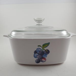 May include: White casserole dish with a clear glass lid. The dish features a painted cluster of blueberries with green leaves. The lid has a clear knob handle. The dish has two small handles on either side.