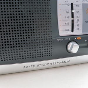 May include: A close-up of an AM/FM weather band radio. The radio has a dark gray speaker grill, a silver base, and a digital clock display. The controls include power, AM/FM band selection, and tuning knobs. The text "AM/FM WEATHER BAND RADIO" is printed on the base.