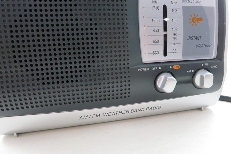 May include: A close-up of an AM/FM weather band radio. The radio has a dark gray speaker grill, a silver base, and a digital clock display. The controls include power, AM/FM band selection, and tuning knobs. The text "AM/FM WEATHER BAND RADIO" is printed on the base.