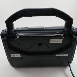 May include: A black portable radio with a retractable antenna and a large, textured handle. The radio has a rectangular shape with a slightly curved top and a series of small buttons and labels. A power cord extends from the bottom.