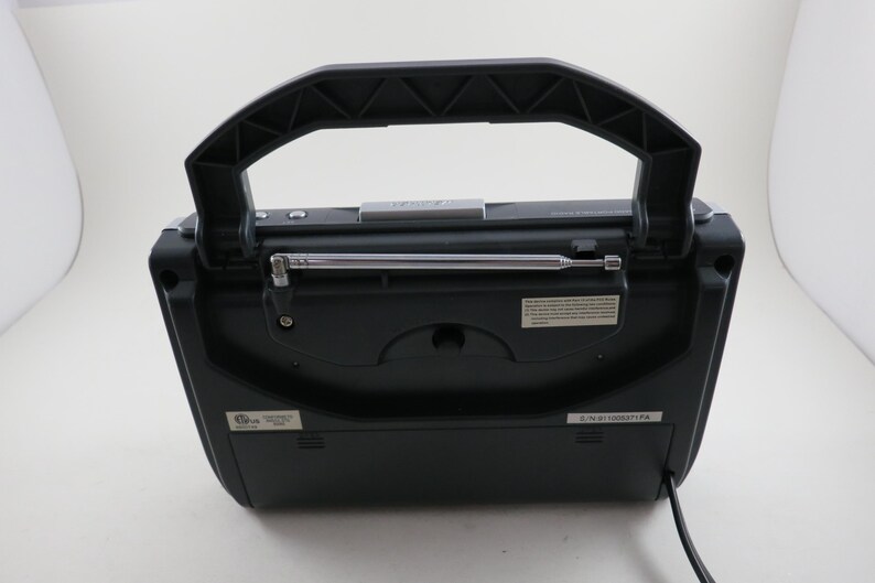 May include: A black portable radio with a retractable antenna and a large, textured handle. The radio has a rectangular shape with a slightly curved top and a series of small buttons and labels. A power cord extends from the bottom.