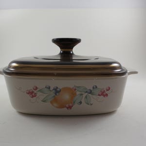May include: A rectangular, off-white casserole dish with a dark brown lid and handle. The dish features a fruit and leaf design in shades of orange, red, green, and blue. The dish has a gold-colored trim and two side handles.