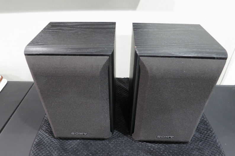 May include: A pair of black Sony bookshelf speakers with a wood grain top finish and grey fabric speaker grilles. Each rectangular speaker unit features the Sony brand name printed in silver lettering at the bottom center of the front panel.