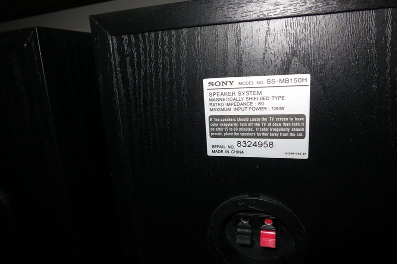 May include: Black speaker cabinet with a textured wood grain finish. A white label displays "SONY" and model information, including "SS-MB150H" and "SPEAKER SYSTEM." The back features a red and black connector.