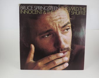 May include: Album cover for Bruce Springsteen's "The Wild, the Innocent & the E Street Shuffle." The cover features a close-up of Springsteen's face, with his hand near his mouth. The album title is printed at the top.