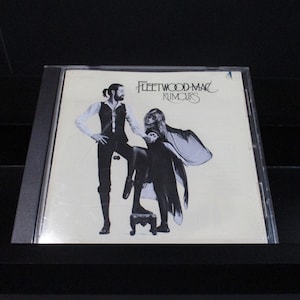 May include: A white CD case with a black and white photo of a man and a woman on the front. The text "Fleetwood Mac" and "Rumours" is printed in black on the front of the case.