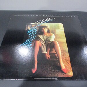 May include: A black vinyl record album cover with the title "Flashdance" in white text. The cover features a woman in a light grey sweater and red shoes sitting on a chair with a patterned rug.