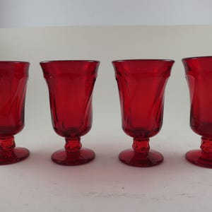May include: Four ruby red stemmed glasses with a decorative swirl design. The glasses have a rounded bowl and a detailed foot. The glasses are arranged in a row against a white background. The glassware is likely for serving beverages.