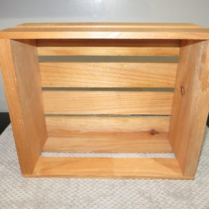 May include: A rectangular wooden crate with an open top and sides constructed from horizontal slats. The crate features a natural light brown wood grain finish with visible knots and smooth edges, providing a sturdy and open storage structure.
