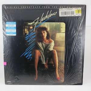 May include: Sealed vinyl record of the "Flashdance" soundtrack. The album cover features a woman in a gray top and red shoes. The title is in a stylized font with blue accents. Includes the text "Original Soundtrack From The Motion Picture".