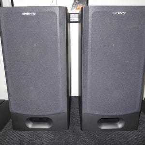 May include: Two dark grey Sony speakers. Each speaker has a rectangular shape with a rounded bottom and a rectangular opening. The Sony logo is visible at the top of each speaker.