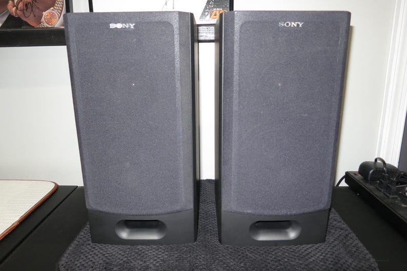 May include: Two dark grey Sony speakers. Each speaker has a rectangular shape with a rounded bottom and a rectangular opening. The Sony logo is visible at the top of each speaker.