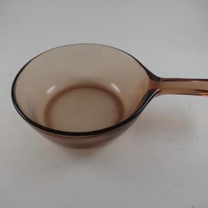 May include: A vintage amber-colored glass saucepan with a handle. The pan has a rounded bowl shape and a dark brown rim. The handle is rectangular and extends from the side of the pan. The saucepan is made of transparent glass.