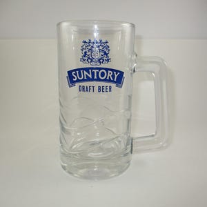 May include: Clear glass beer mug with a handle. The mug features the blue text "SUNTORY DRAFT BEER" and a crest. The glass has a textured design on the lower portion of the mug.