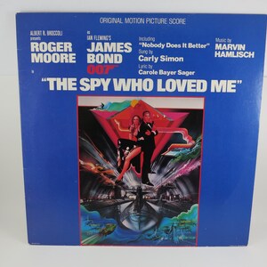 May include: A vintage vinyl record album cover for "The Spy Who Loved Me" featuring Roger Moore as James Bond. The cover is predominantly blue with text and an illustration of a man and woman. The album includes the song "Nobody Does It Better".