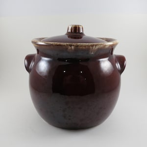 May include: A dark brown ceramic bean pot with a lid. The pot has two handles and a glossy finish. The lid has a small knob on top. The pot is approximately 8 inches tall.