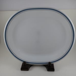 May include: An oval white serving platter featuring a simple blue band design along the rim. The platter is smooth and glossy, with a clean, minimalist aesthetic. It is displayed upright on a dark brown stand, highlighting its shape and color.