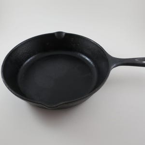 May include: A black cast iron skillet with a long handle and a small pouring spout. The skillet is round with a flat cooking surface and a loop at the end of the handle for hanging. The pan is made of heavy-duty metal.
