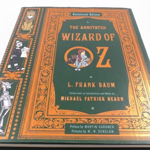 May include: A hardcover book titled The Annotated Wizard of Oz, Centennial Edition by L. Frank Baum. The dark green cover features orange decorative borders, illustrations of the Cowardly Lion, Scarecrow, and Tin Woodman, and a central graphic of Dorothy and Toto.