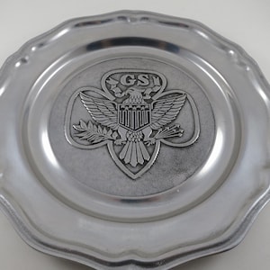 May include: A silver-colored plate with a scalloped edge. The center features a raised emblem of an eagle with outstretched wings, a shield, and the letters "GS". A decorative item.