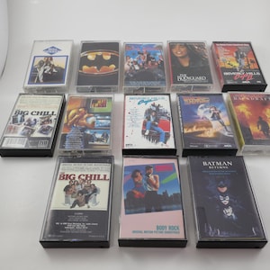 May include: A collection of 12 VHS tapes featuring various movie titles, including "The Big Chill", "Batman Returns", "Beverly Hills Cop", "The Breakfast Club", "Say Anything", "Backdraft", "The Bonfire of the Vanities", "Predator", "The Fly", "Twins", "The Jewel of the Nile", and "Romancing the Stone".