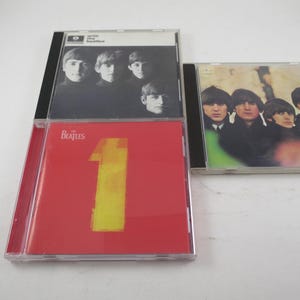 May include: Three Beatles CD albums. One album has a red cover with a large yellow number 1. Another album has a black and white photo of the band members. The third album has a green and white cover with a photo of the band.