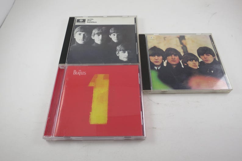 May include: Three Beatles CD albums. One album has a red cover with a large yellow number 1. Another album has a black and white photo of the band members. The third album has a green and white cover with a photo of the band.