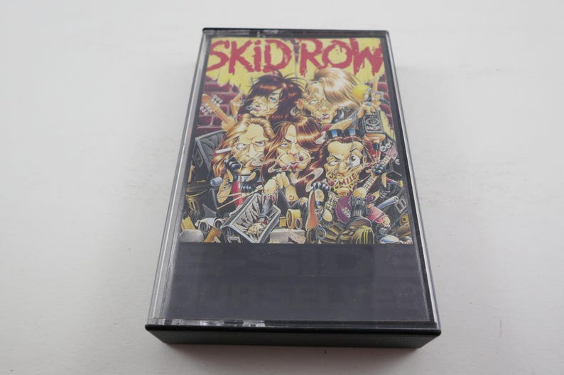 May include: A cassette tape with a black case and a colourful image of a band called "Skid Row". The image features five cartoon figures with exaggerated features, including a guitarist, a drummer, and a singer.