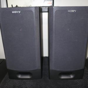 May include: A pair of black Sony speakers with dark grey fabric fronts. Each speaker has a rectangular shape with a slightly angled top and a rectangular port at the bottom. The Sony logo is visible on the top.