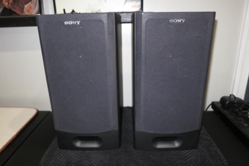 May include: A pair of black Sony speakers with dark grey fabric fronts. Each speaker has a rectangular shape with a slightly angled top and a rectangular port at the bottom. The Sony logo is visible on the top.