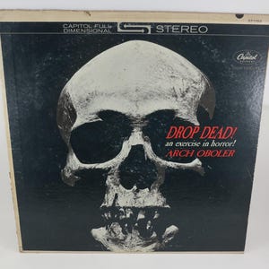 May include: Vintage vinyl record album cover for "Drop Dead!" by Arch Oboler. The cover features a large, detailed skull graphic in shades of white and gray against a black background. The album title and artist name are in red.