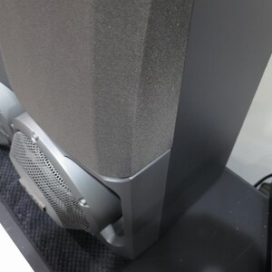 May include: Two black speakers with gray mesh grilles and dark gray foam fronts. The speakers are on a black surface, with a power strip and other electronics visible in the background. The speakers are likely part of a home audio system.
