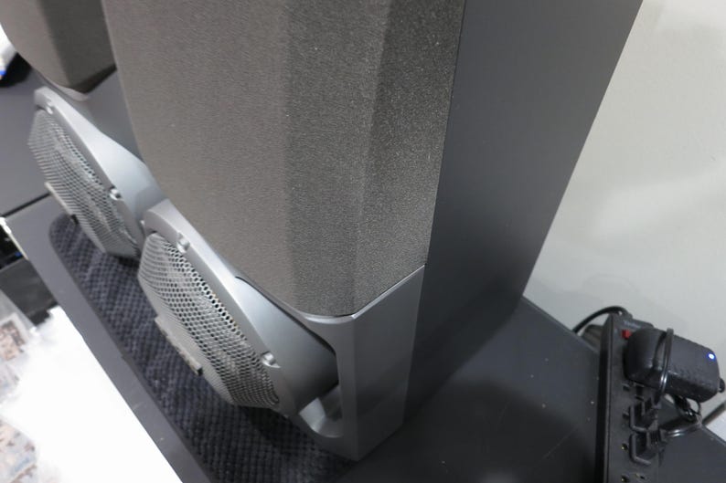 May include: Two black speakers with gray mesh grilles and dark gray foam fronts. The speakers are on a black surface, with a power strip and other electronics visible in the background. The speakers are likely part of a home audio system.