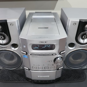 May include: A silver Sony stereo system featuring a central unit with a digital display showing 042, multiple control buttons, and a large volume dial, flanked by two matching silver speakers with black accents and visible circular speaker cones.