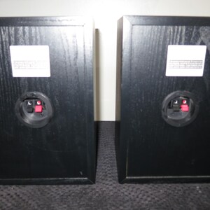 May include: Two black rectangular speakers with a wood grain finish. Each speaker has a white label and a circular connector with red and black terminals. The speakers are positioned side by side.