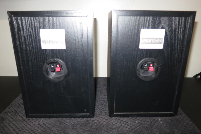 May include: Two black rectangular speakers with a wood grain finish. Each speaker has a white label and a circular connector with red and black terminals. The speakers are positioned side by side.