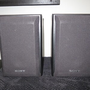 May include: A pair of rectangular black Sony speakers featuring dark grey fabric grilles covering the front panels. Each speaker has the Sony brand name printed in silver lettering at the bottom center of the front face, with a solid black frame.